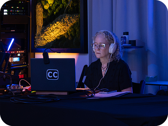 A captioner wearing headphones works at a laptop displaying the CC (closed captions) logo in a dimly lit event venue.