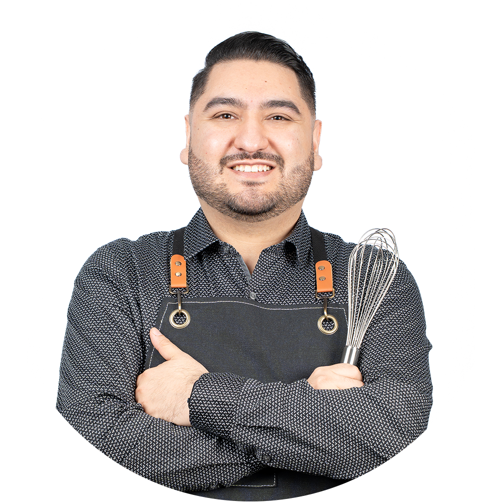 Oscar smiles with his arms crossed, wearing a kitchen apron and holding a whisk.